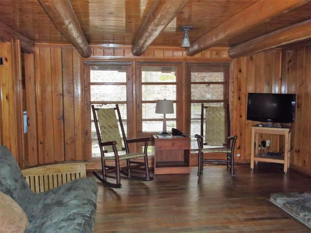 A view of the living room area in cabin 11 including a couch, tv and two rocking chairs in front of three large exterior windows